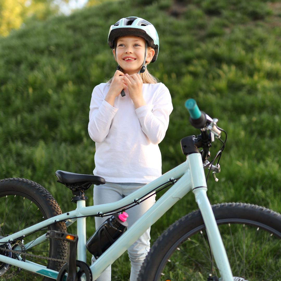 A young child is standing outdoors next to a bicycle, smiling while fastening their helmet strap. She is wearing a white long-sleeve shirt and light-colored pants, and appears to be getting ready for a bike ride. The bicycle is light blue and the background is a grassy outdoor area.