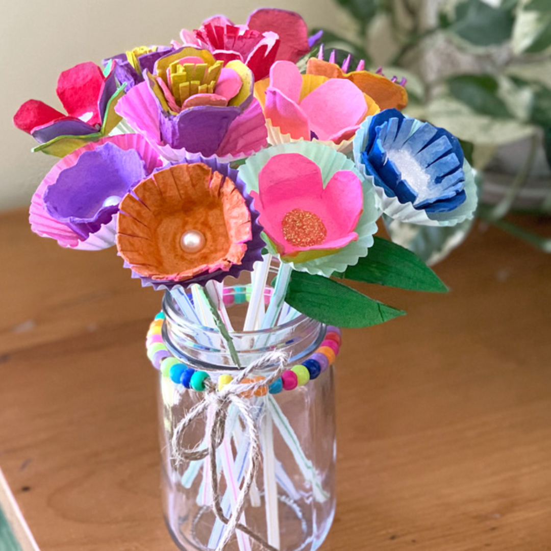 Photo of a bouquet of craft flowers made of egg carton pieces and painted bright colors sitting inside a clear glass vase on a wooden table.