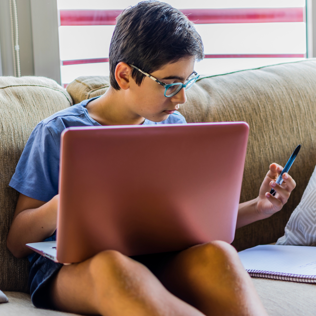 A teen wearing glasses is sitting on a couch at home, focused on a laptop placed on their lap. They’re holding a pen in one hand and looking down at a notebook beside them, suggesting they’re studying or doing homework.