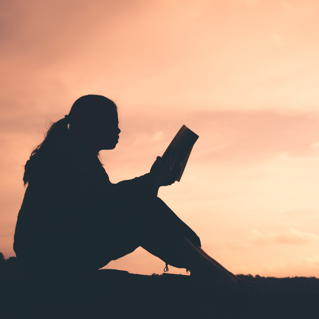 A shadow figure of a person sits on the ground outside reading a book with a soft pink sky in the background.