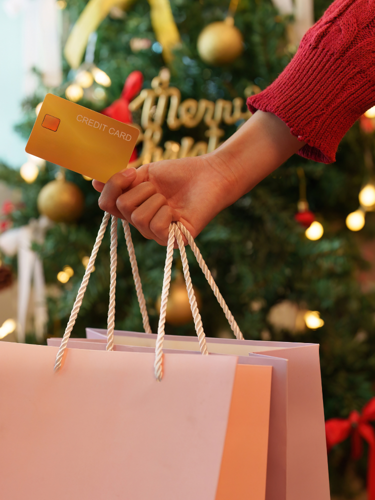 Person holding gift bags and credit card in front of Christmas tree.