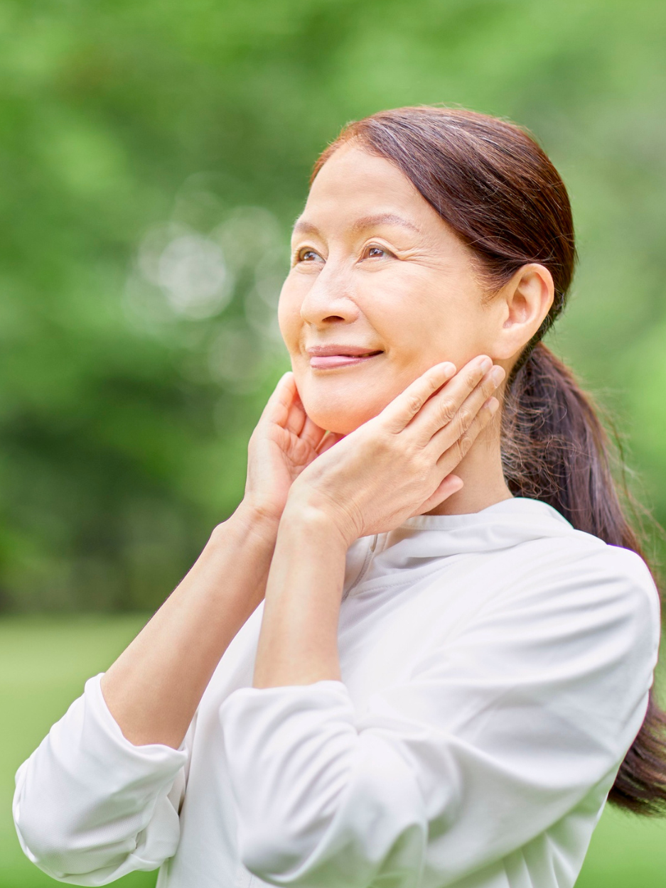 A woman standing outdoors in a green park setting, gently holding her hands to her neck and jawline while looking slightly upward with a calm, content expression. She is wearing a light gray long-sleeve top.