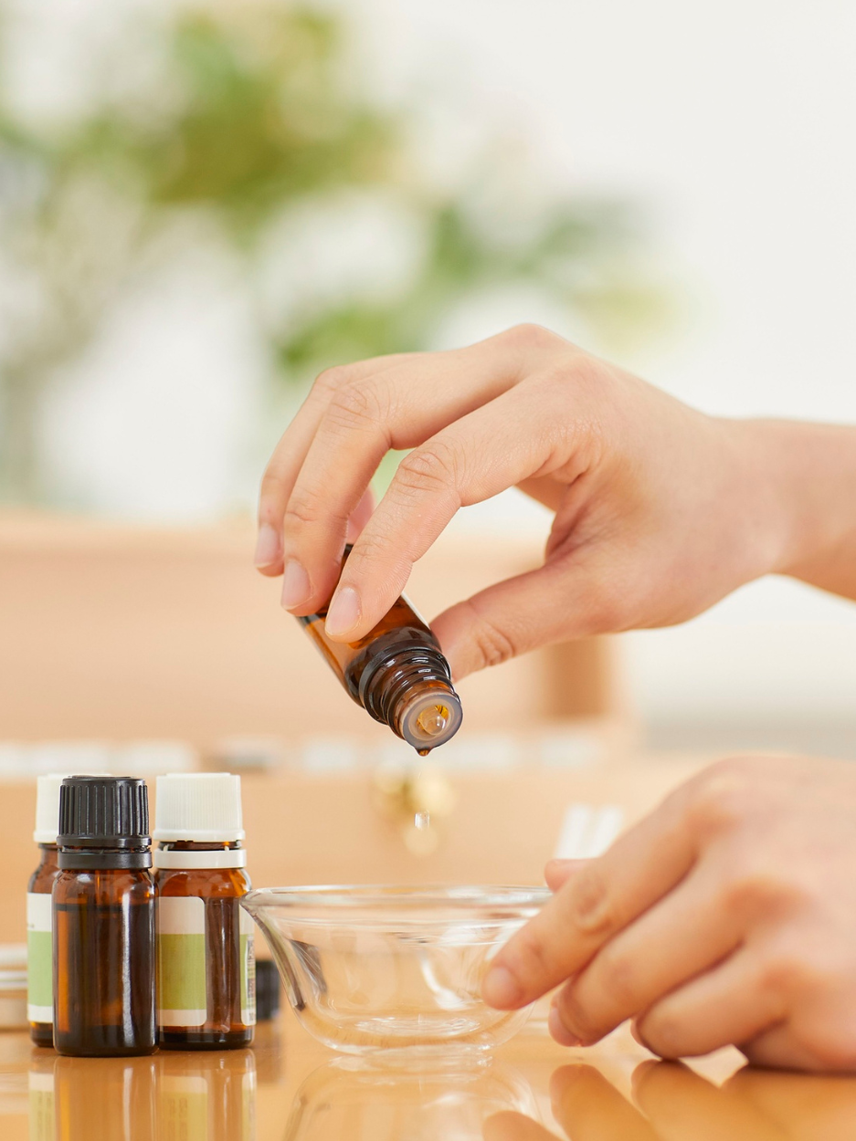 The image shows a pair of hands pouring a small glass amber bottle of fragrance oil into a small clear glass bowl on top of a table. There are three more small amber glass fragrance vials sitting on the table.