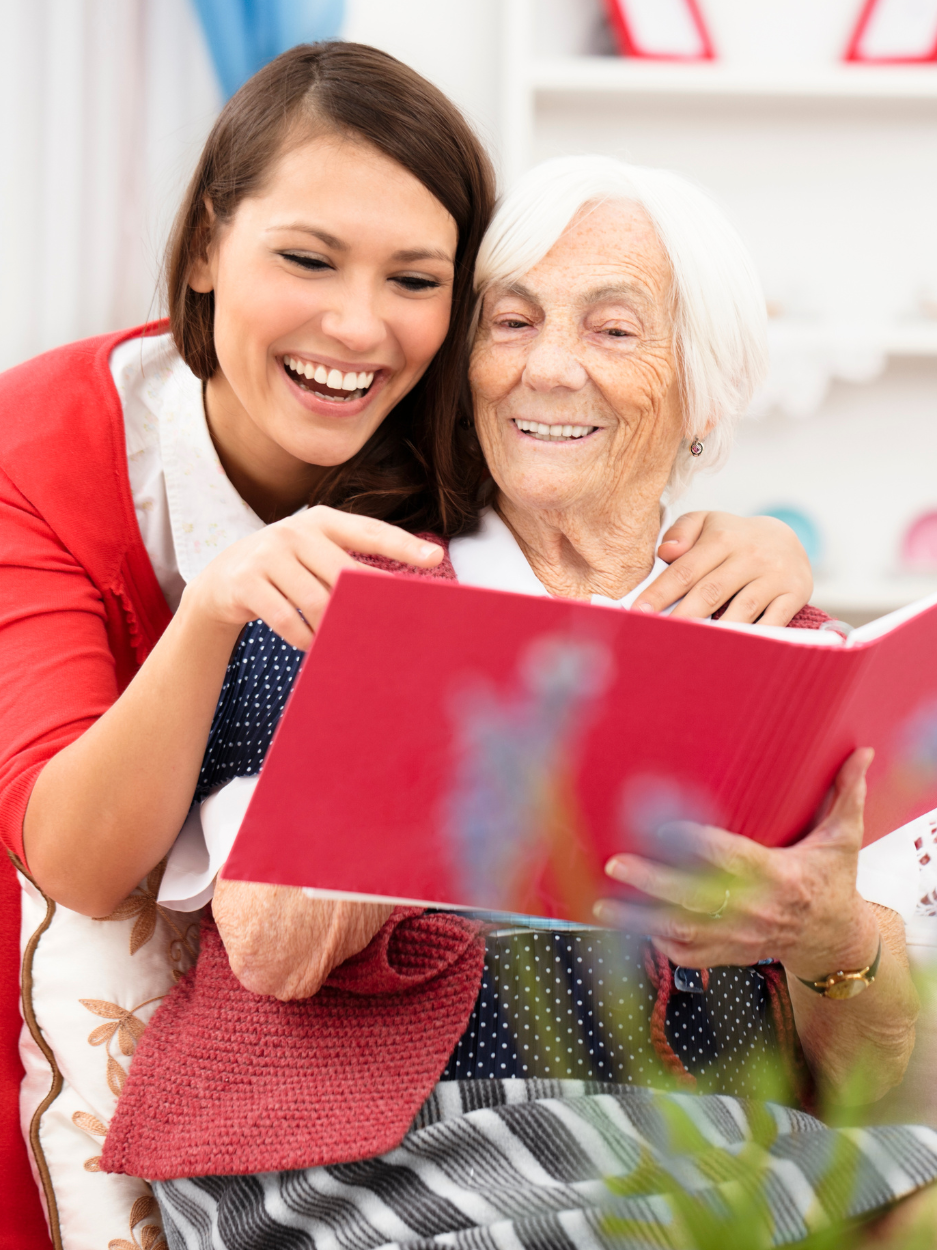 A smiling younger woman sits close beside an elderly woman with white hair, wrapping an arm around her shoulders as they look at and read a red book together. Both appear happy and engaged. They are indoors in a bright setting with soft-focus shelves in the background.