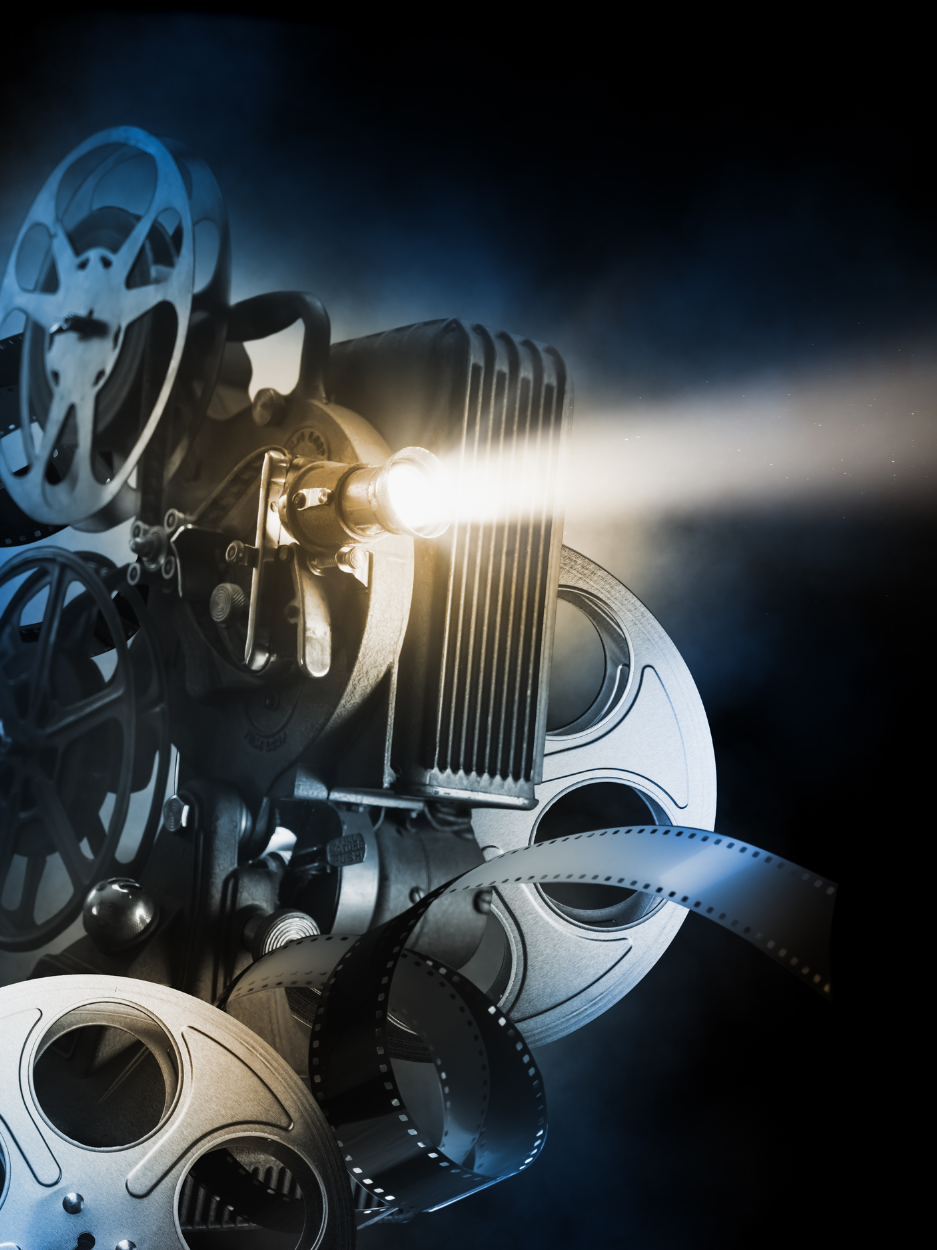 Image of a film projector with film reels in a dark room that illuminates a film being screened off the image.