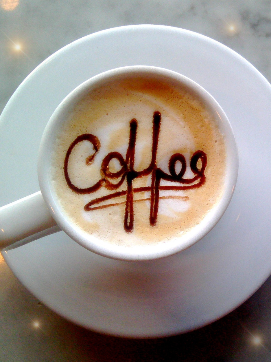 photograph of a white mug and saucer filled with a latte with the word coffee written in the foam