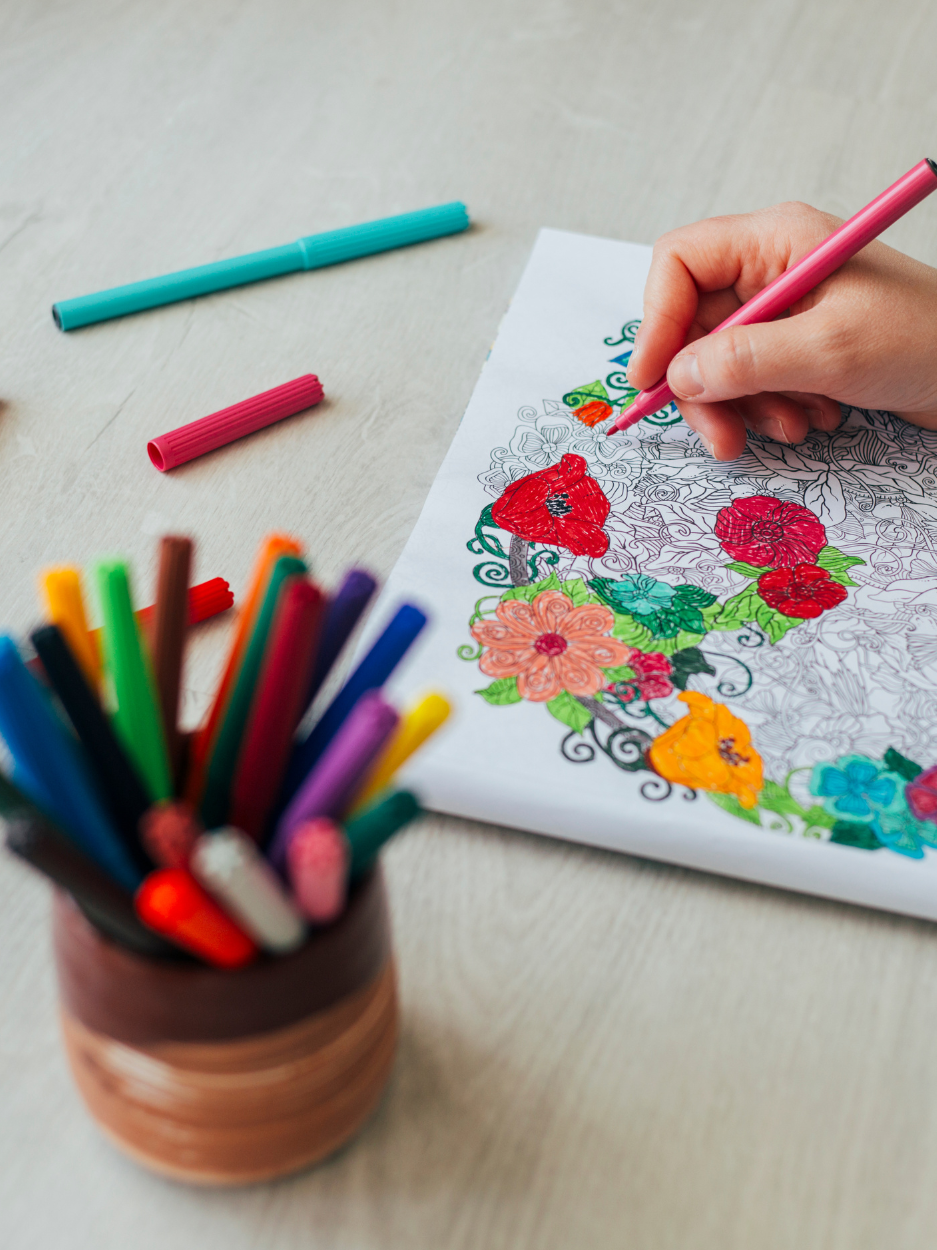 The image shows a close-up of a person coloring a detailed floral design on a sheet of paper. They are using a pink marker to fill in parts of the illustration. On the table, there are several colorful markers scattered around, along with a container holding many more markers in different colors.