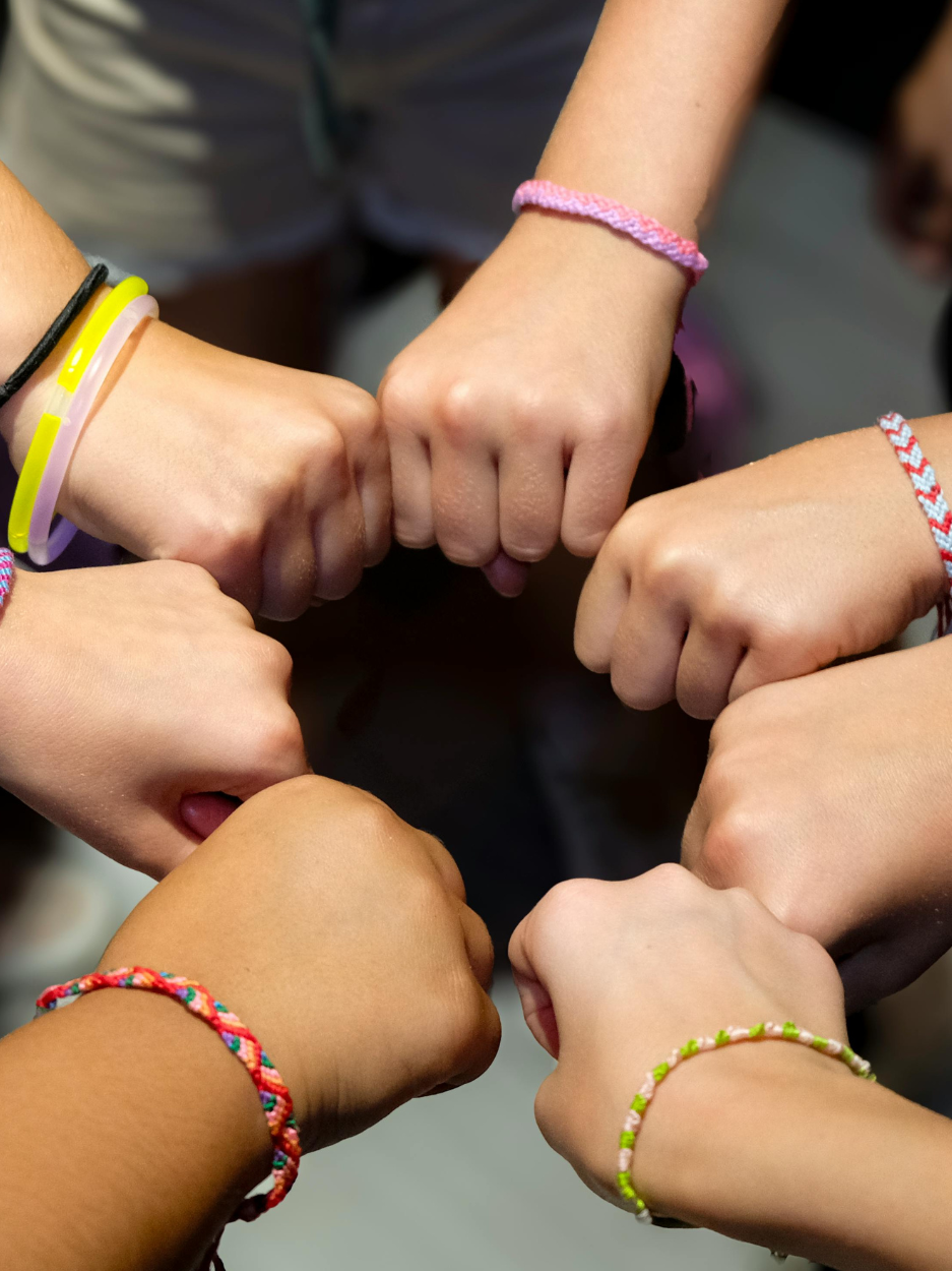 Six people forming a circle with their fists touching in the center, each wearing colorful friendship bracelets and wristbands.