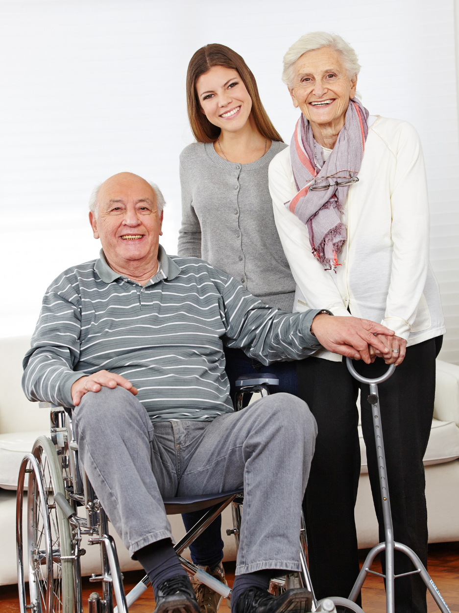 An elderly man sitting in a wheelchair smiles at the camera while holding hands with an elderly woman standing beside him, who is using a cane. A younger woman stands behind them with her arms around their shoulders.