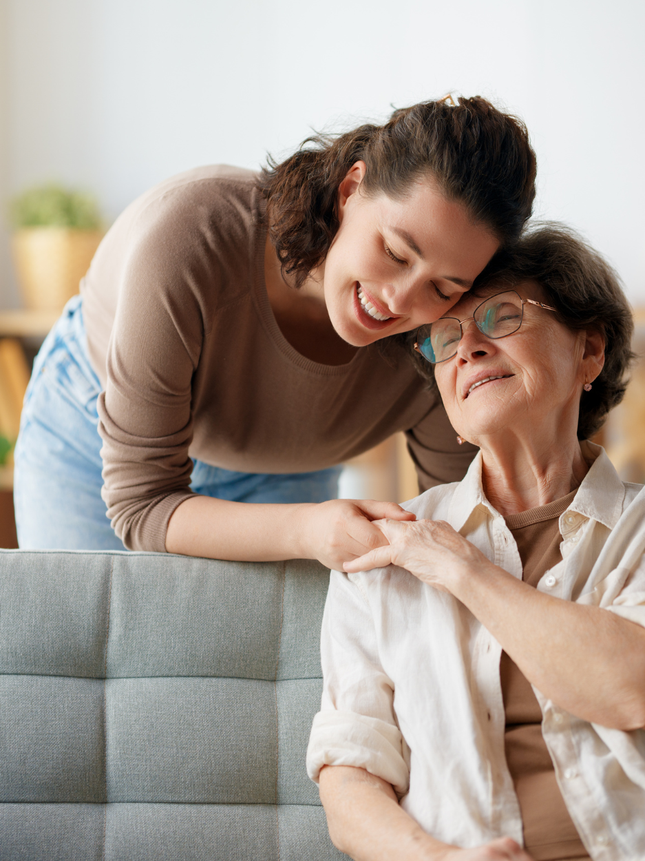 A young woman leans over the back of a couch, smiling warmly as she gently rests her head against an older woman’s head. The older woman, wearing glasses and a light button-up shirt, smiles with her eyes closed while holding the younger woman’s hand. The setting appears to be a living room.