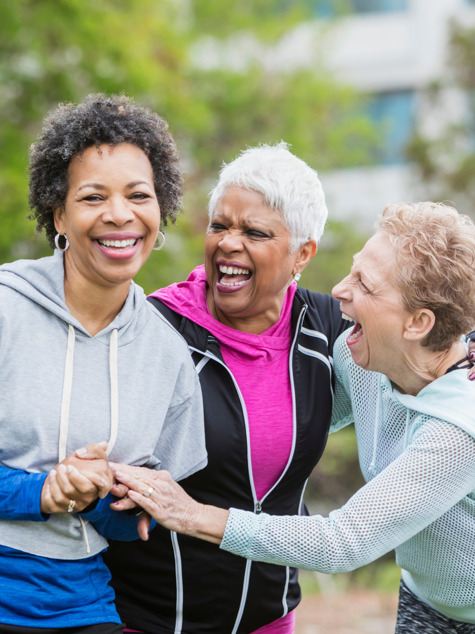 Three older women standing close together outdoors, smiling and laughing. They are dressed in casual athletic wear. They have their arms around each other in a friendly, supportive pose, with greenery and a building softly blurred in the background.