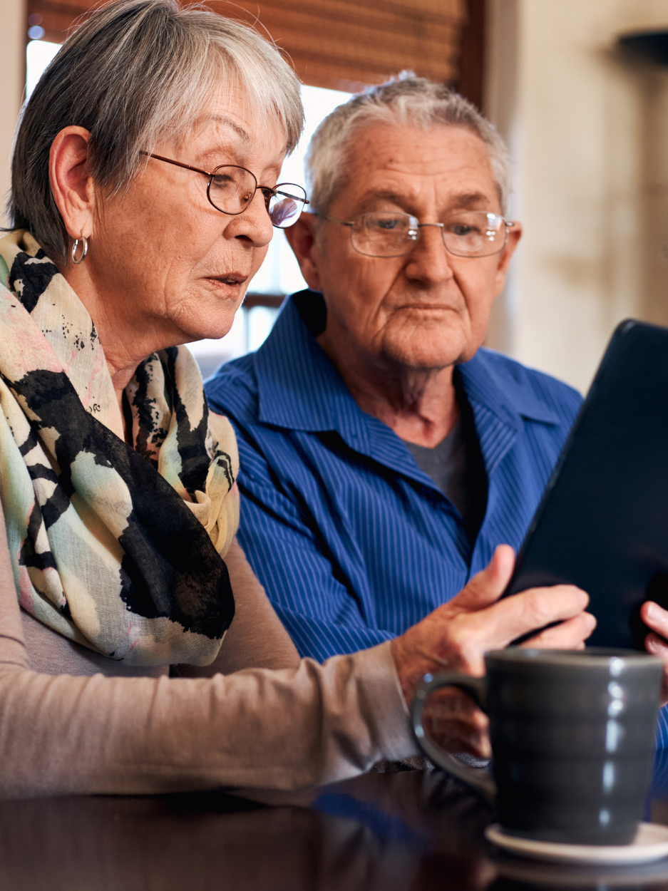 An elderly woman and an elderly man sit side by side at a table, looking intently at a tablet the man is holding. Both wear glasses, and a coffee mug sits on the table in front of them.