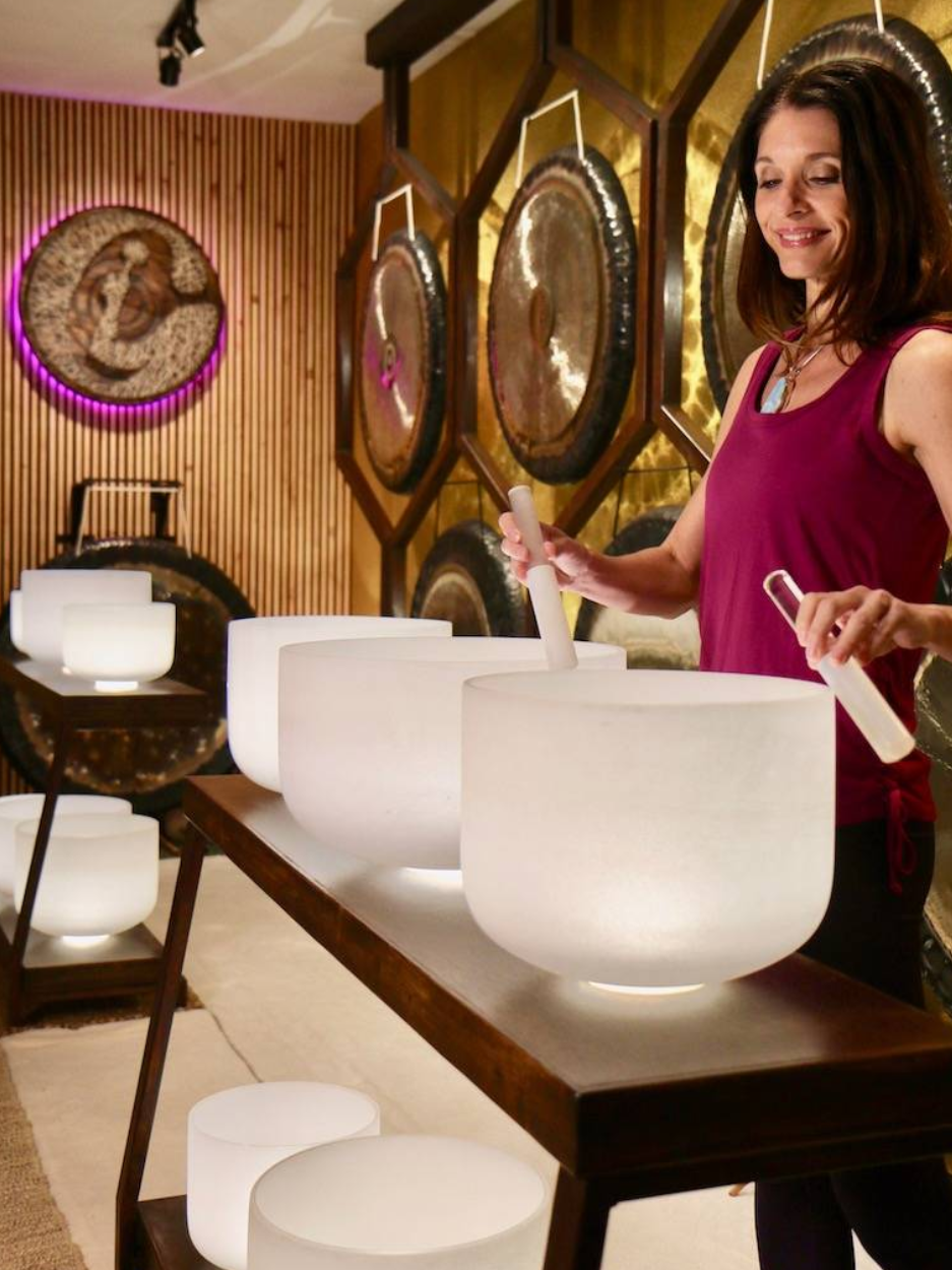 A woman is standing in a room playing crystal sound bowls. She is holding a white mallet in each hand and gently striking or circling the rims of large frosted white bowls arranged on a wooden table. Behind her, several large bronze gongs hang on a wooden wall with vertical slats.