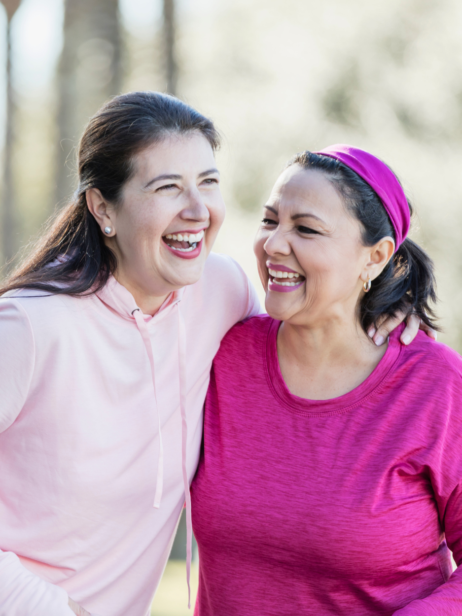 Two women standing outdoors with their arms around each other, laughing and smiling. One woman is wearing a light pink hoodie, the other is wearing a bright pink athletic top. The background is softly blurred with trees. Their expressions convey a happy moment.