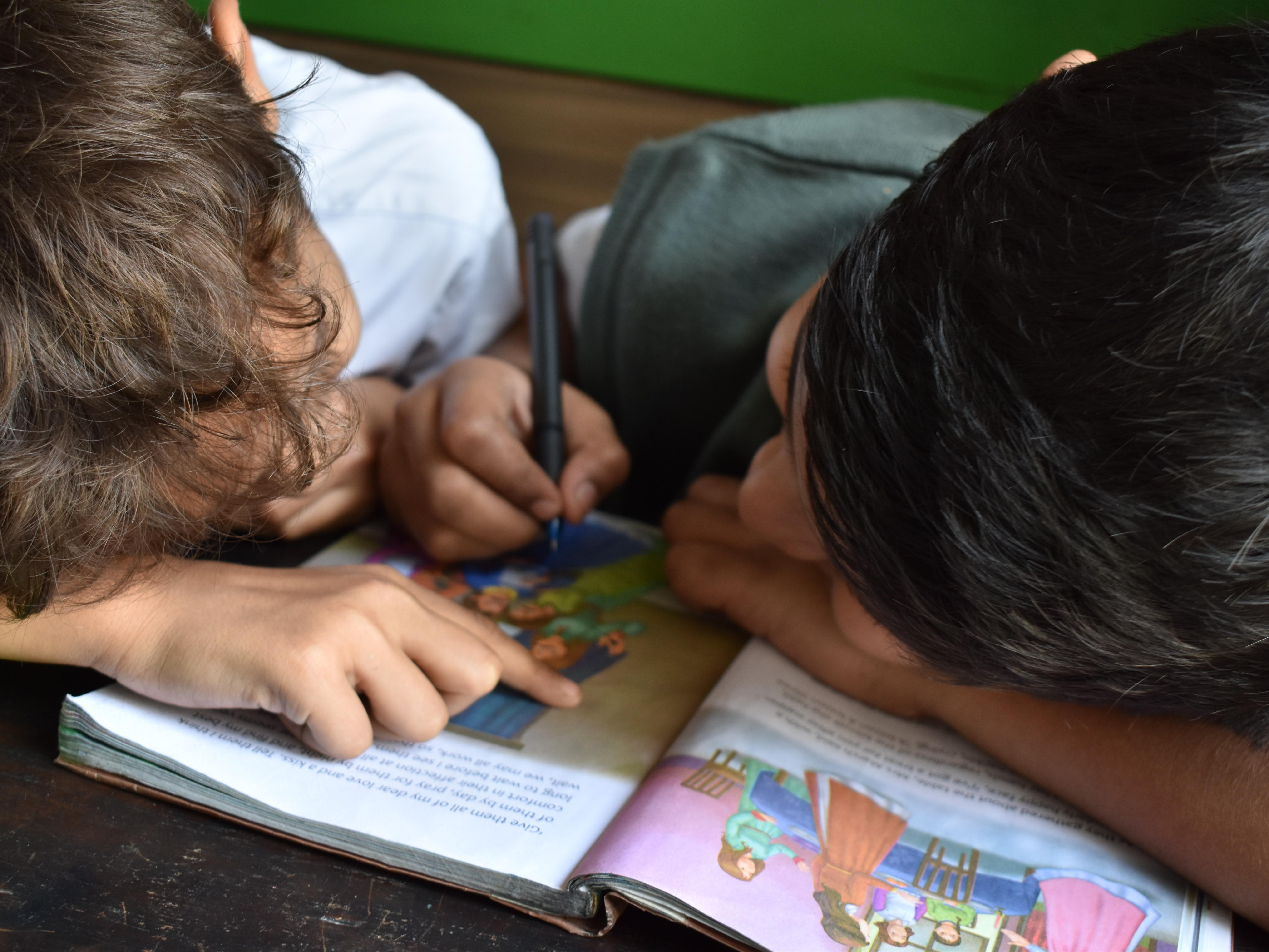 two children leaning over a book together, one using a pen