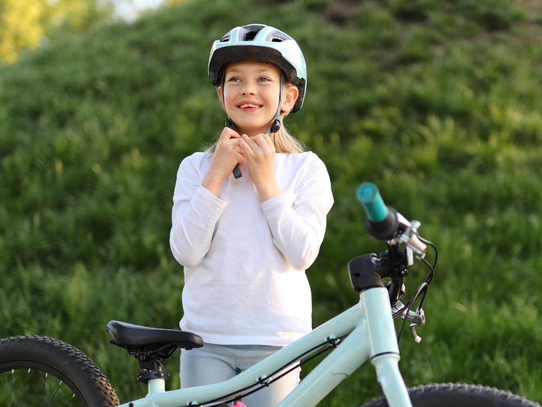 A young child is standing outdoors next to a bicycle, smiling while fastening their helmet strap. She is wearing a white long-sleeve shirt and light-colored pants, and appears to be getting ready for a bike ride. The bicycle is light blue and the background is a grassy outdoor area.