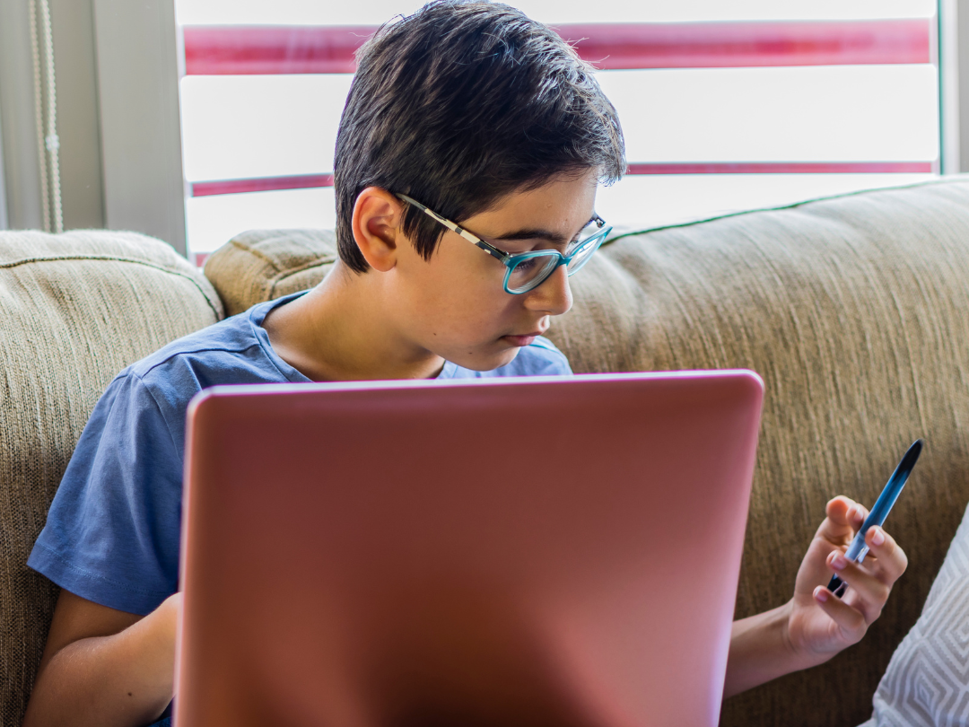 A teen wearing glasses is sitting on a couch at home, focused on a laptop placed on their lap. They’re holding a pen in one hand and looking down at a notebook beside them, suggesting they’re studying or doing homework.